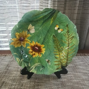 May include: A green ceramic plate with a leafy design featuring yellow flowers, butterflies, and a bee. The plate has a scalloped edge and is on a black stand.