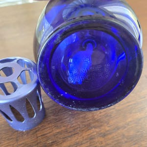 Pu&ograve; includere: Una bottiglia di vetro blu cobalto con la scritta "LAMPE BERGER PARIS" in rilievo sul fondo. Un diffusore in metallo argentato &egrave; posizionato accanto alla bottiglia. La bottiglia ha una forma arrotondata.