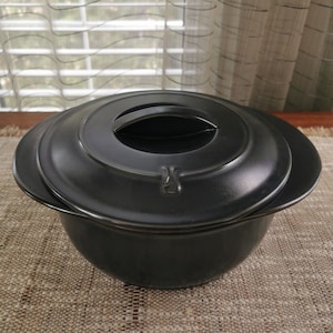 Puede incluir: Plato para hornear de cerámica negra con tapa y asa en la tapa. El plato está sobre una base tejida de color marrón.