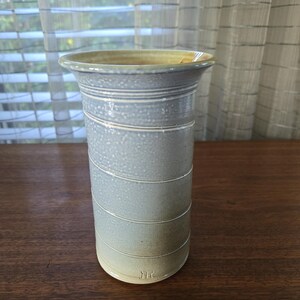 Pode incluir: Um vaso de cerâmica feito à mão com um gradiente azul claro e verde. O vaso cilíndrico apresenta linhas gravadas horizontais e uma borda alargada. A base tem uma marca estampada. Aproximadamente 20 cm de altura.