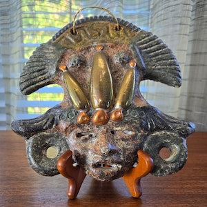 May include: A ceramic mask with a human face, painted in brown, green, and gold. The mask has a crown of gold and copper accents and is displayed on a wooden stand.