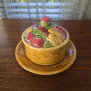 May include: A ceramic butter dish with a basket weave design. The lid is adorned with a variety of colorful, sculpted fruits, including strawberries, bananas, and an apple. The dish sits on a matching plate.