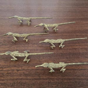 May include: Six small, brass-coloured roadrunner figurines are arranged on a dark wood surface. The figurines are detailed with outstretched legs and long beaks, capturing the bird's running posture. The roadrunners are approximately 7.6 cm long.