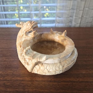 Puede incluir: Un cenicero de dragón tallado, de color beige. El dragón está enrollado alrededor de un cuenco circular, con la cabeza levantada y la boca abierta. El cenicero está hecho de piedra o cerámica de color claro, y los detalles del dragón están finamente tallados.