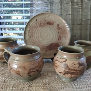 Puede incluir: Conjunto de cuatro tazas de cerámica marrón y verde con un plato de cerámica marrón y blanco. Las tazas tienen una forma redondeada y un asa. El plato tiene una forma circular y un diseño marrón y blanco.