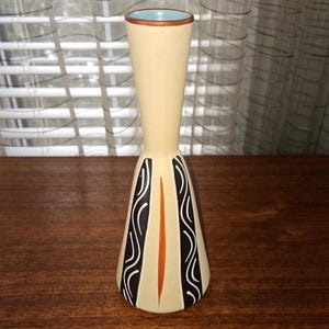 Może przedstawiać: Kremowy ceramiczny wazon z rozszerzoną podstawą i wąską szyjką. Wazon posiada czarno-białe wzory fal, pomarańczowy pionowy pas i brązową obwódkę. Wnętrze jest jasnoniebieskie.