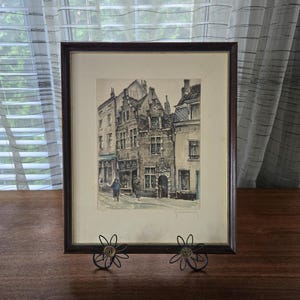May include: A framed black and white print of a street scene in a European city. The print depicts a narrow street with buildings on both sides. The buildings are tall and have many windows. The street is paved with cobblestones and there are people walking on the street.