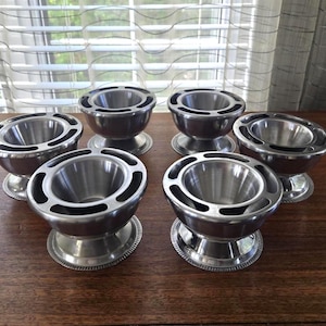 May include: A set of six silver-toned metal egg cups arranged in two rows. Each cup features a smooth, rounded bowl set upon a circular pedestal base with a decorative, textured rim and a wide, flat top edge with evenly spaced cutouts.