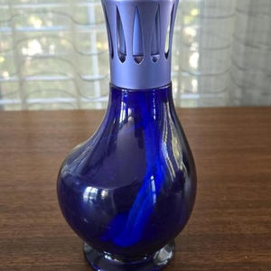 Pu&ograve; includere: Una lampada profumata in vetro blu scuro con una parte superiore in metallo color argento. La lampada ha un corpo arrotondato e una base svasata. La parte superiore ha un design decorativo con fessure verticali. La lampada &egrave; progettata per diffondere oli profumati.
