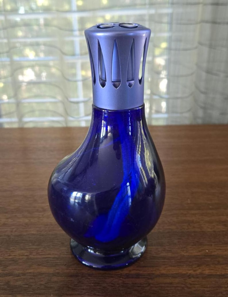 Pu&ograve; includere: Una lampada profumata in vetro blu scuro con una parte superiore in metallo color argento. La lampada ha un corpo arrotondato e una base svasata. La parte superiore ha un design decorativo con fessure verticali. La lampada &egrave; progettata per diffondere oli profumati.