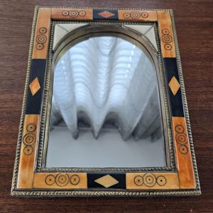 Puede incluir: Un pequeño espejo rectangular con un marco ornamentado. El marco está hecho de madera y hueso, y presenta un patrón geométrico de círculos, diamantes y cuadrados. El espejo está rodeado por un fino borde de metal dorado.