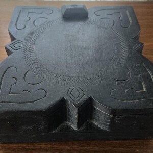 May include: A dark, carved wooden box with a hinged lid. The box features intricate floral and geometric designs, including a central oval with a leaf pattern. The box is rectangular with a decorative clasp.