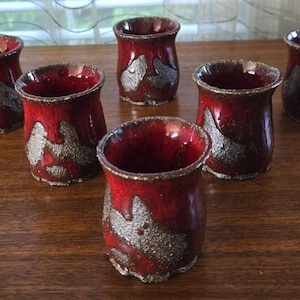 May include: Six handmade ceramic shot glasses. The glasses are a deep red with a metallic silver design. The interior of the glasses is a glossy red. The glasses are arranged on a dark wooden surface.