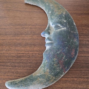 May include: A crescent moon wall hanging featuring a sculpted human face in profile. The surface has a weathered, distressed finish with mottled shades of dark green, brown, and hints of red, giving it a textured, aged appearance throughout.