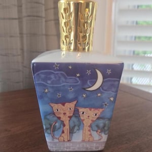 May include: A square-shaped ceramic fragrance lamp featuring a night sky illustration with a crescent moon, stars, and two cats sitting on a brick wall. The lamp is topped with a gold-coloured metal cap with a cut-out leaf pattern design.