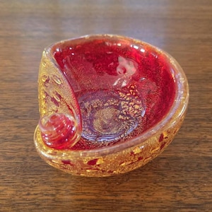 Puede incluir: Un cuenco decorativo de cristal con interior rojo y motas doradas. El cuenco tiene un diseño único en espiral con un pequeño acento redondeado. El cristal tiene un acabado brillante. El cuenco está sobre una superficie de madera.