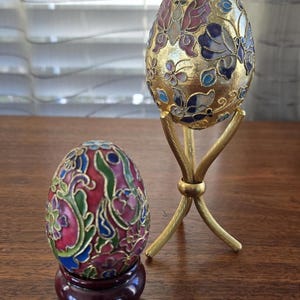 May include: Two decorative eggs with intricate floral and butterfly designs. The eggs are painted in vibrant colours, including gold, red, blue, green, and pink. The larger egg is on a gold stand, while the smaller egg is on a dark wood stand.
