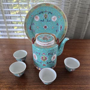 Könnte beinhalten: Ein türkisfarbenes Teeservice mit passendem Teller. Das Set enthält eine Teekanne, vier kleine Tassen und einen Teller. Die Teekanne und der Teller weisen ein Blumen- und geometrisches Design mit roten und weißen Akzenten auf. Das Teeservice wird auf einer Holzoberfläche präsentiert.