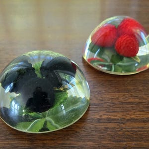 May include: Two clear glass paperweights, each containing a different type of fruit. One paperweight contains red raspberries, while the other contains blackberries.