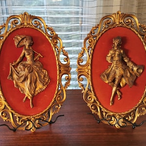 May include: Two gold-toned framed wall sculptures with ornate frames. Each frame features a three-dimensional figure of a person in a vintage style dress and suit. The figures are set against a red velvet background.
