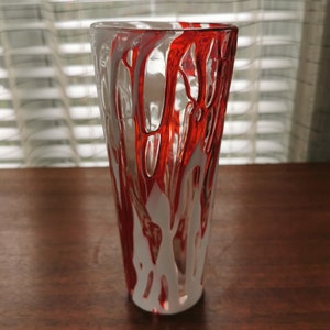 May include: A clear glass vase with red and white paint dripping down the sides. The vase is tall and slender, with a rounded bottom.