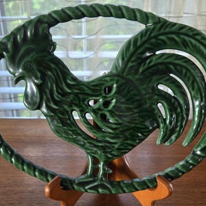 May include: Green ceramic rooster-shaped hot pad with a rope-like design around the edge. The hot pad is on a wooden stand.