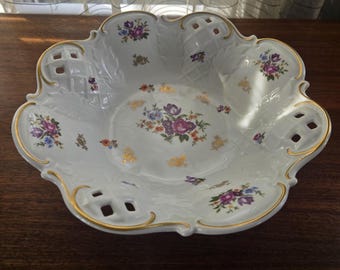 Puede incluir: Bol de porcelana blanca con borde festoneado y ribete dorado. El bol presenta un estampado floral en tonos morado, rosa y azul. El bol tiene un diseño calado decorativo y está sobre una superficie de madera oscura.