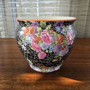May include: A ceramic planter with a black crackle glaze, featuring a colourful floral design. The flowers are in shades of pink, orange, purple, and blue. The rim has an orange accent.