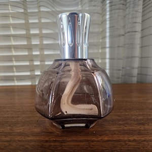 Puede incluir: Una lámpara de fragancia decorativa de vidrio teñido de púrpura con una parte superior de metal de color plateado. La lámpara tiene una mecha visible dentro del recipiente de vidrio. La lámpara está sobre una superficie de madera.