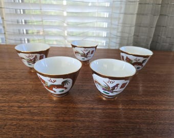 May include: Five white teacups with gold trim and colorful dragon designs. The cups are arranged in a semi-circle on a wooden surface.