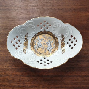 Op de afbeelding: Witte ovale schaal van porselein met een gouden rand en een delicaat opengewerkt ontwerp. Het midden heeft een rond gouden ontwerp met twee engelen in een bloemrijke omgeving.