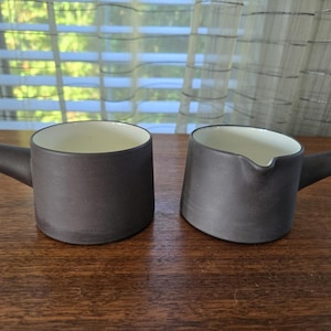 Può includere: Due tazze in ceramica marrone scuro con interni bianchi e manici. Una è una tazza standard, l'altra ha un beccuccio. Le tazze sono su una superficie di legno.