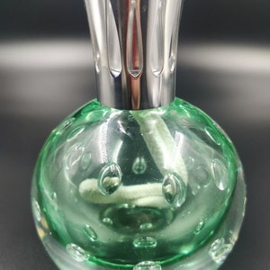 Op de afbeelding: Een groene glazen oliebrander met een zilveren metalen deksel en een witte lont. Het glas heeft een getextureerd oppervlak met kleine bubbels.