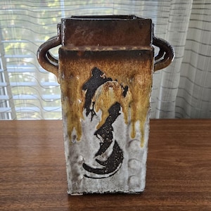 May include: Rectangular ceramic vase with a brown top and handles, featuring a dripped glaze effect in shades of brown and yellow. The front displays a black abstract design on a white background. A decorative piece.
