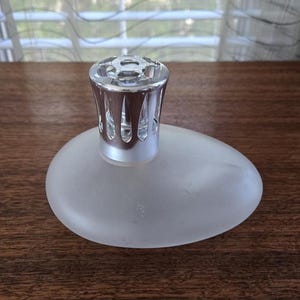 Puede incluir: Un difusor de fragancia de vidrio esmerilado con una tapa de metal plateado. El difusor tiene forma ovalada y se apoya sobre una superficie de madera. La tapa plateada tiene un diseño decorativo y abierto.