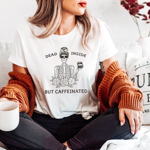 May include: A white t-shirt with a black and white illustration of a skeleton holding a coffee cup. The text on the shirt reads "Dead Inside But Caffeinated".