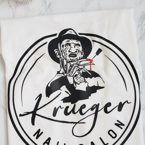 Puede incluir: Camiseta blanca con un gr&aacute;fico en blanco y negro de Freddy Krueger y el texto "Krueger Nail Salon" en un dise&ntilde;o circular. El dise&ntilde;o incluye un sombrero negro y un cuchillo ensangrentado. Una peque&ntilde;a planta es visible en la esquina superior izquierda.