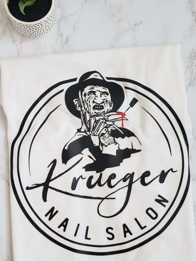 Puede incluir: Camiseta blanca con un gr&aacute;fico en blanco y negro de Freddy Krueger y el texto "Krueger Nail Salon" en un dise&ntilde;o circular. El dise&ntilde;o incluye un sombrero negro y un cuchillo ensangrentado. Una peque&ntilde;a planta es visible en la esquina superior izquierda.