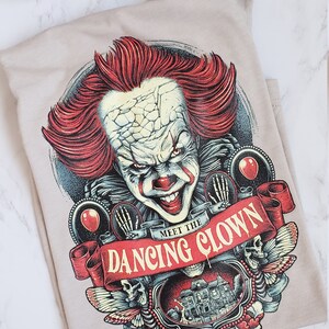 Op de afbeelding: Een beige T-shirt met een grafische print van een enge clown met rood haar en een rode neus. De clown wordt omringd door schedels, vlinders en een banner met de tekst "Meet the Dancing Clown".