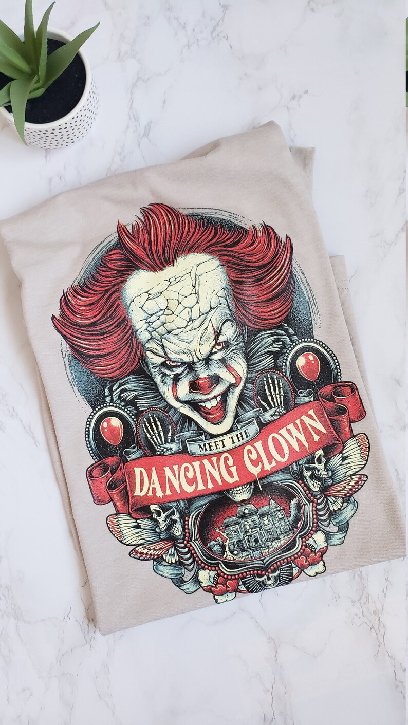 Op de afbeelding: Een beige T-shirt met een grafische print van een enge clown met rood haar en een rode neus. De clown wordt omringd door schedels, vlinders en een banner met de tekst "Meet the Dancing Clown".