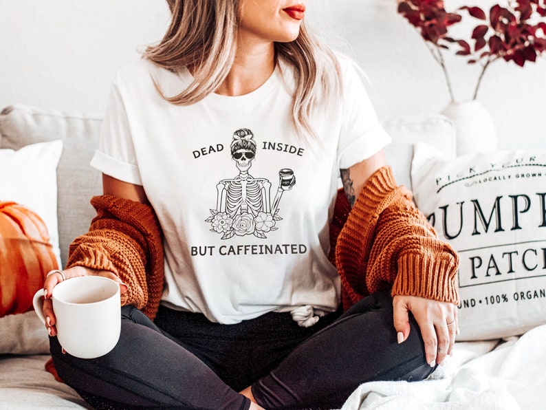 May include: A white t-shirt with a black and white illustration of a skeleton holding a coffee cup. The text on the shirt reads "Dead Inside But Caffeinated".