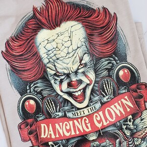 Op de afbeelding: Een beige T-shirt met een grafische print van een enge clown met rood haar en een dreigende glimlach. De tekst "Meet the Dancing Clown" is gedrukt op een rood lint.