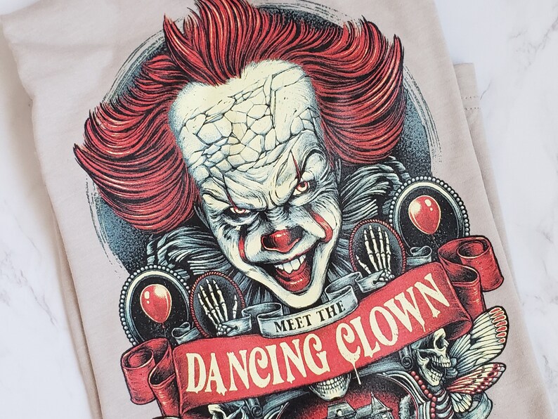 Op de afbeelding: Een beige T-shirt met een grafische print van een enge clown met rood haar en een dreigende glimlach. De tekst "Meet the Dancing Clown" is gedrukt op een rood lint.