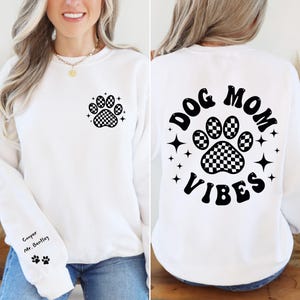 May include: White sweatshirt with a black checkered paw print on the left chest and "DOG MOM VIBES" with a black checkered paw print in a circle on the back. The text is in a retro font.