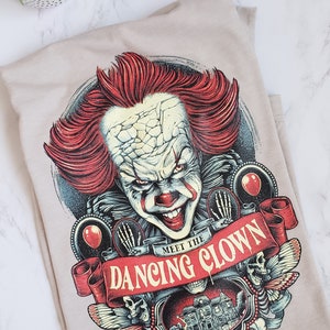 Op de afbeelding: Een beige T-shirt met een grafische print van een enge clown met rood haar en een dreigende glimlach. De grafiek bevat ook een rode banner met de tekst "Meet the Dancing Clown".