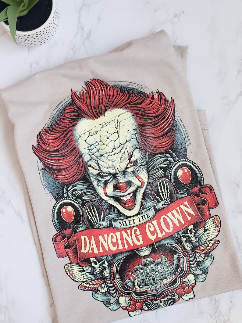 Op de afbeelding: Een beige T-shirt met een grafische print van een enge clown met rood haar en een dreigende glimlach. De grafiek bevat ook een rode banner met de tekst "Meet the Dancing Clown".