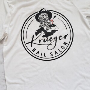 Puede incluir: Camiseta blanca con un gr&aacute;fico negro y rojo de un personaje con sombrero y guante con cuchillas, rodeado por el texto "Krueger Nail Salon". El dise&ntilde;o est&aacute; centrado en la camiseta.