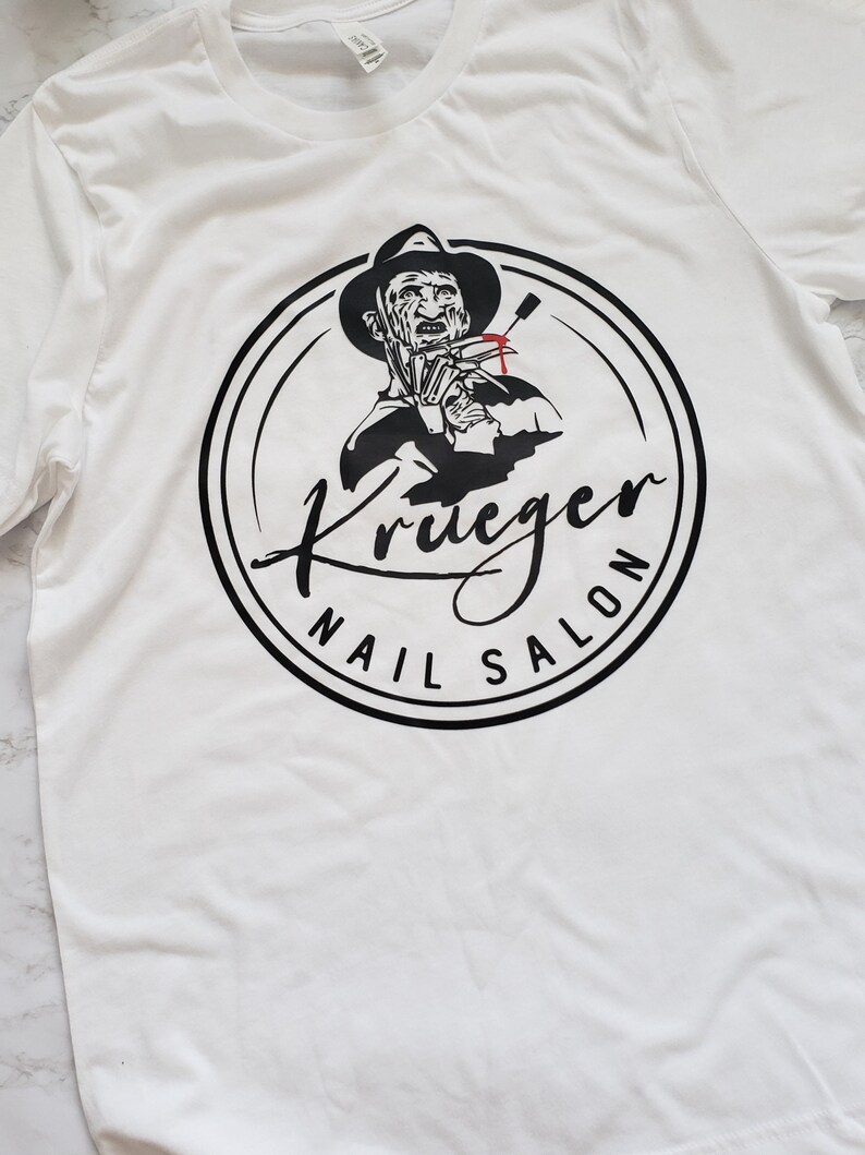 Puede incluir: Camiseta blanca con un gr&aacute;fico negro y rojo de un personaje con sombrero y guante con cuchillas, rodeado por el texto "Krueger Nail Salon". El dise&ntilde;o est&aacute; centrado en la camiseta.