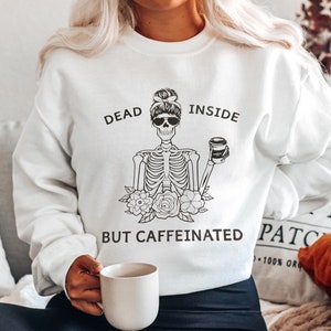 May include: White sweatshirt with a black and white illustration of a skeleton holding a coffee cup. The text "Dead Inside But Caffeinated" is printed on the sweatshirt.