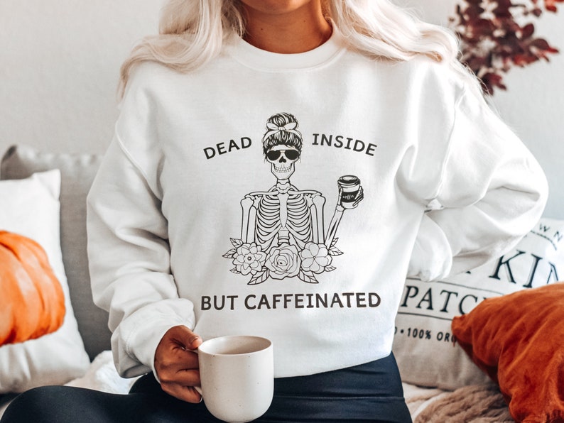 May include: White sweatshirt with a black and white illustration of a skeleton holding a coffee cup. The text "Dead Inside But Caffeinated" is printed on the sweatshirt.